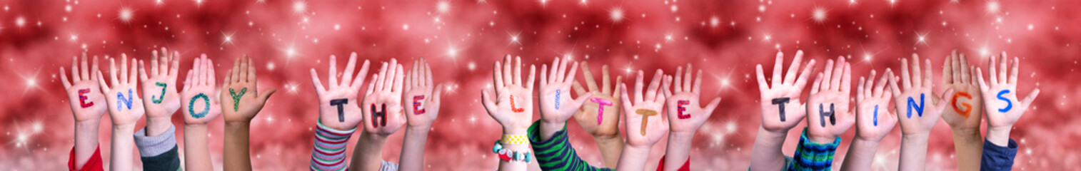 Children Hands Building Colorful English Word Enjoy The Little Things. Red Snowy Christmas Winter Background With Snowflakes And Sparkling Lights