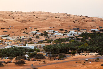 A small village with houses and green trees is located in the middle of a large sand dunes in the desert.