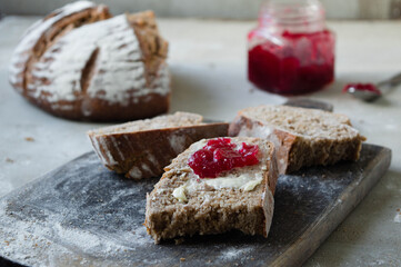 Pieces of homemade whole grain wheat and rye bread dusted with cherry jam and butter on the vintage wooden pan, glass jar with cherry jam