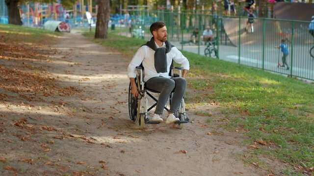 Young portrait serious handsome disabled man rides in wheelchairat at sunlight in park. Paralysis disability positive handicapped. Slow motion