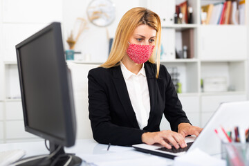 Young businesswoman in colored medical mask working with laptop and documents in office