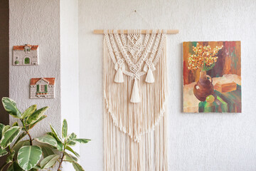 Macrame wallhanding. Decoration for the interior. Interior design of  living room with beige beautiful macrame, canvas painting, green plants and elegant accessories. Concept of cozy home decor.