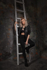 Beautiful blonde girl with exquisite makeup and long legs in a black bra jacket and trousers near an old wall with a wooden staircase