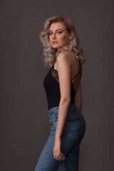 Fototapeta premium Beautiful blonde girl with exquisite makeup and long legs in a black T-shirt and jeans on a gray background