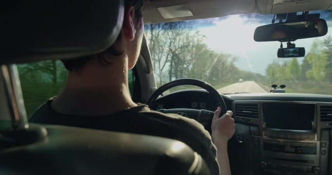 Rear View to the young man drives a car. Teenager drives the car while sunset.  Young man driving the car.  Teen boy  driving car. Sun's rays shine through the windshield of the car. Real time.