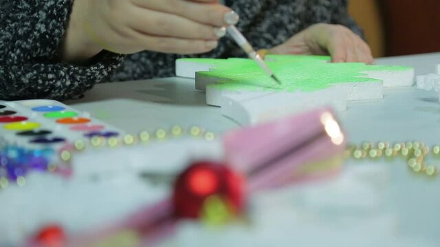 On a winter evening, a young woman makes home decorations for Christmas by painting a Christmas tree figurine with green watercolor