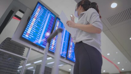 Asian Young woman wear mask standing in front of airport flight time schedule screen looking checking counter on travel itinerary paper, at airport terminal, lady and suitcase luggage low angle view