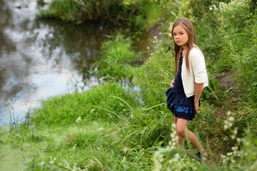 Beautiful blonde girl child in a black dress and white sweater near a small river
