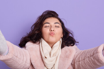 Young joyful brunette girl holding taking selfie self portrait of herself standing against lilac background, keeps eyes closed, expresses positive, wears warm faux fur coat and scarf.