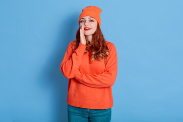Portrait of cute, trendy, charming, positive woman holding palm near mouth, screaming news information, calling someone isolated on blue background, wearing orange jumper, cap and jeans.
