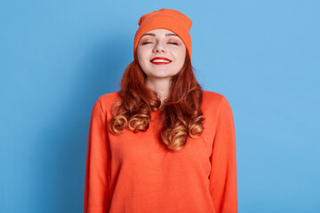 Horizontal shot of winsome young female with red hair, keeps eyes closed, imagines pleasant moment, wearing new bought sweater and warm hat, isolated on blue background.