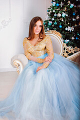 Fototapeta premium Young and happy woman in long party dress sitting near Christmas tree with Christmas toys in festive studio