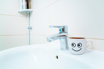 chromed metal faucet for hot and cold water,  cup with smile face for toothbrushes  in a modern bathroom