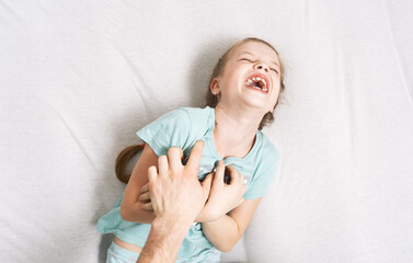Family time: a little girl lies on the bed and laughs cheerfully because of the tickling of her...
