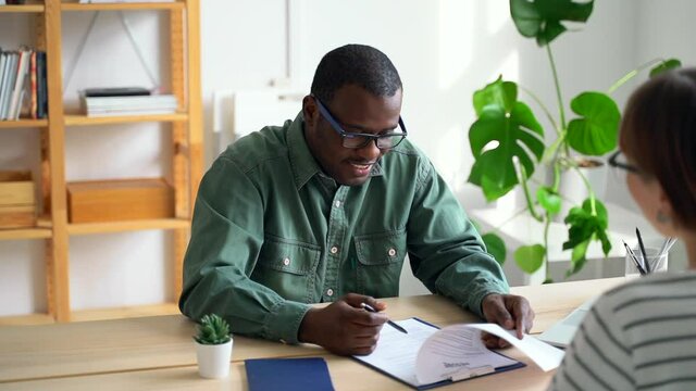 HR employee talking to female job seeker and looking resume at table in modern office spbas. African American man interviews woman and reads document with smile while sitting at desk in bright room