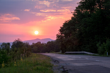 Country road at sunrise. The sun rises against the background of the mountains. Green grass and a road stretching into the distance.