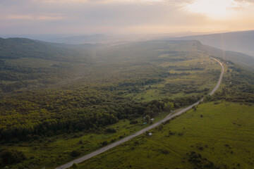 Early morning in the mountains. Morning sun and haze in the valley against the background of mountains. Green fields and meadows. The road leaving into the distance. Adygea, Russia