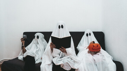 Trio of halloween ghosts sitting in the sofa reading, drinking wine and playing with pumpkin