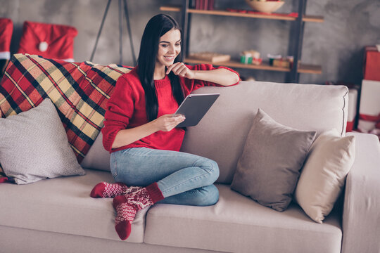 Photo of pretty young girl sit sofa hold tablet hand chin look screen wear red pullover jeans socks in decorated living room indoors