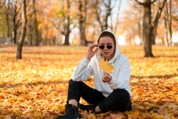 Woman in a white sweater with a hood sits on ground in the park and holding a leaf in her hands