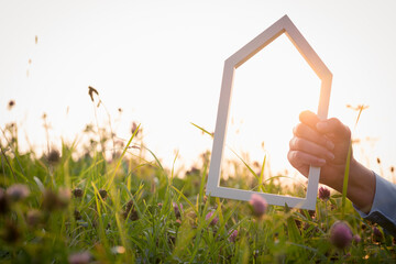 A woman's hand holding a white wooden house on a meadow during a beautiful sunset atmosphere