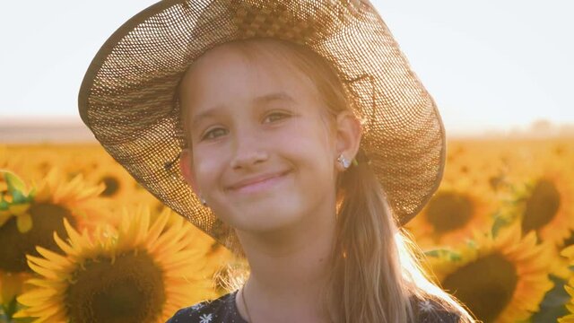 A happy little girl in the hat is smiling at the field of sunflowers at sunset time.