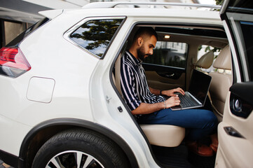 Fototapeta na wymiar Successful arab man wear in striped shirt and sunglasses pose inside white suv car with laptop in hands.