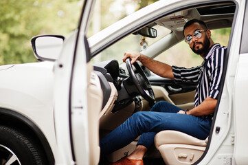 Successful arab man wear in striped shirt and sunglasses pose behind the wheel of  his white suv car. Stylish arabian men in transport.