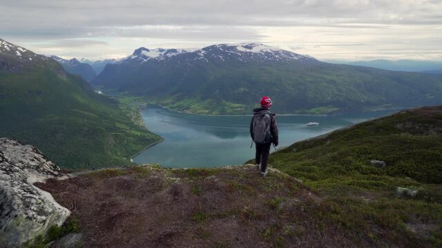 Child hiking on mountain peak overlooking Norwegian fjord and lake. Cruiser ship in instance. Summer time Norway nature view. Smooth gimbal shot at Nordfjord and Oldevatnet natural enviroment