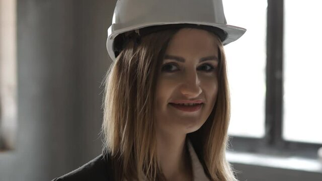 Portrait of an attractive young woman engineer or worker in a uniform and protective white helmet, smiling and looking at camera at construction site