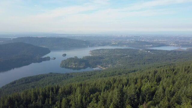 Vancouver Indian Arm and Deep Cove from high above over forest. Drone Aerial shot of green pine trees, blue water, buildings, skyline in the distance. Summer morning light in British Columbia Canada