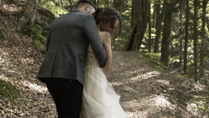 Among the autumn forest with brown and green leaves on the trail, a guy in a jacket and black pants with a beard catches up with a smiling and happy girl in a white lush dress and hugs her and kisses