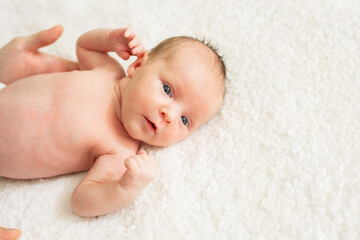 Cute naked newborn baby with open eyes, lying on a bed. A parent holds the legs of a newborn baby.