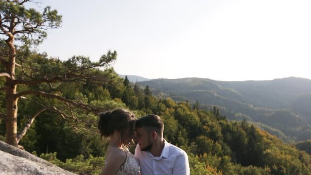 Young married couple sitting high in the mountains on a large rock at sunset with clear blue sky, guy with a beard stroking hand of a girl with a beautiful hairstyle white elegant dress on his stomach