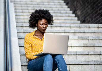 Fototapeta na wymiar Young African American woman in casual clothing, sitting on the stairway and using her laptop computer
