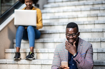 Young African American man sitting and looking at his i phone, casually dressed and smiling while responding on text message