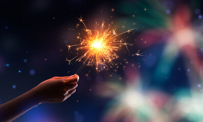 Hand holding a burning sparkler against fireworks background - Powered by Adobe