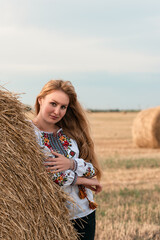 Naklejka na ściany i meble Beautiful ukrainian young woman in a field with wheat bales in embroidered shirt
