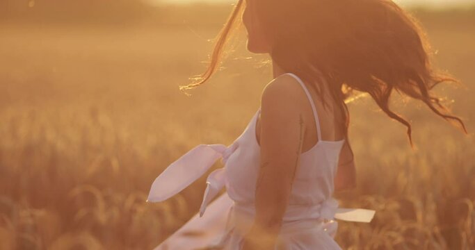 Stock footage of a carefree brunette woman in white summer dress dancing, twisting and rotating in golden wheat field in bright sunlight.