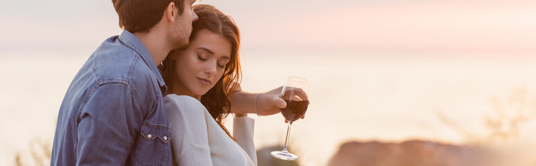 Fototapeta premium Website header of man hugging woman while holding glass of wine on beach at sunset