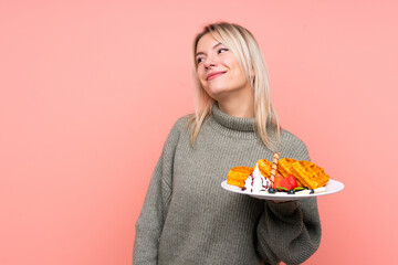 Young blonde woman holding waffles over isolated pink background looking up while smiling