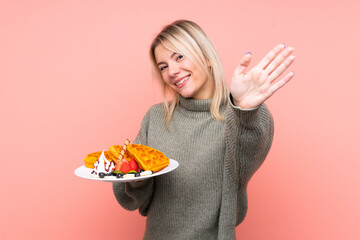 Young blonde woman holding waffles over isolated pink background saluting with hand with happy expression