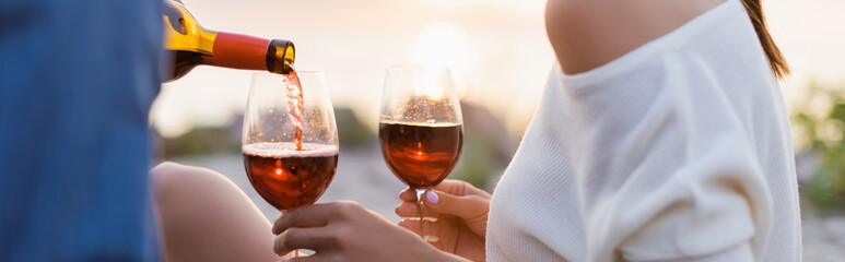 Panoramic shot of man pouring wine in glasses near girlfriend on beach