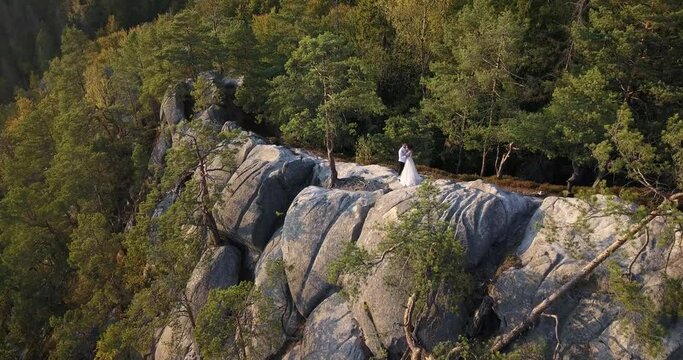 In the mountains on a stone ridge of large boulders among the autumn green and yellow pine trees on a warm day at sunset is a couple in love in white clothes. Quadcopter flying over rocks to them