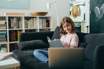 Fototapeta na wymiar young girl having online school lessons, sitting on sofa in her home