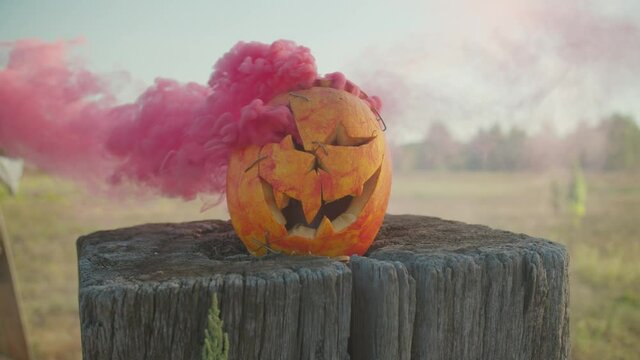 Close-up of spooky smashed carved halloween pumpkin with colorful red smoke coming out of holes lying on wooden stump over rural background.