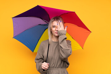 Young blonde woman holding an umbrella over isolated yellow wall covering a eye by hand