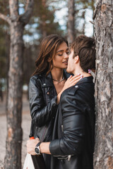 Selective focus of woman kissing boyfriend in leather jacket with bottle of wine in forest