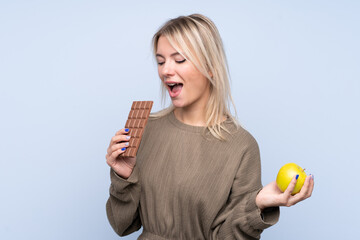 Young blonde woman over isolated blue background taking a chocolate tablet in one hand and an apple...