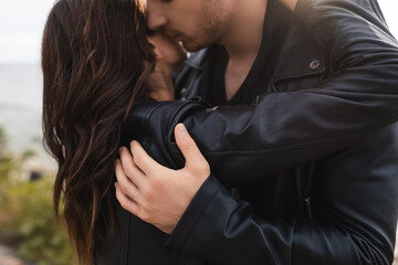 Man kissing and embracing brunette girlfriend in leather jacket on beach